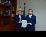 À frente, dois homens posando para a foto segurando uma cartilha de boas ações. Ao fundo, uma parede na cor branca, três bandeiras hasteadas e uma estante com livros à esquerda. 
