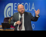 Homem vestido com terno escuro, camisa branca e gravata de cor marrom. Com a mão direita, segura o microfone e gesticula com a mão esquerda. A parede ao fundo da imagem na cor azul. 