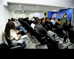 Foto de homens e mulheres sentados em um auditório, que estão assistindo a uma mulher que está em pé e falando ao microfone, em frente a uma parede azul, onde está escrito Escola Judicial. A mulher tem cabelos compridos e escuros e está vestindo um macacão verde. 