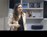Foto de uma mulher branca, de cabelos longos, usando óculos, vestindo roupas nas cores preta e bege.  