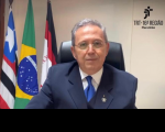 Homem sentado em uma cadeira preta, usa óculos de grau com lentes arredondadas, um terno azul escuro, uma camisa branca e uma gravata azul. Ao fundo, uma parede na cor branca, três bandeiras hasteadas.