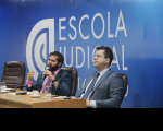 Foto de dois homens sentados à mesa de honra de evento da Escola Judicial, um deles falando ao microfone. Atrás, parede azul onde se lê Escola Judicial.