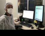 Imagem de um homem usando touca, máscara e avental descartáveis na cor branca. Ele está sentado numa cadeira branca, em frente a uma mesa onde estão duas telas de computador, um teclado e outros equipamentos.