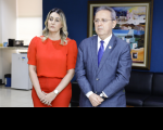 Foto de duas pessoas, um homem e uma mulher, ambos com as mãos cruzadas à frente de seus corpos. A  mulher, ao lado esquerdo, vestida com roupas de cor vermelha. O homem, vestido com terno escuro, camisa branca e gravata azul.