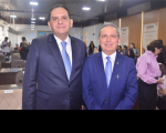 Dois homens em um plenário posando para foto, ambos vestidos com terno e gravata azul e camisa branca. O homem da direita usa óculos de grau. Ao fundo, algumas pessoas sentadas e outras em pé.