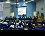 Foto de várias pessoas (homens e mulheres) sentadas num auditório participando de seminário; no destaque autoridades da solenidade de abertura. Na parede central do auditório, um projetor com as informações do evento. À direita, o brasão do Judiciário na parte superior da parede.
