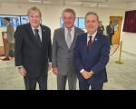 Três homens posando para foto, ambos vestidos com ternos. O homem da esquerda veste terno preto, o do meio veste terno cinza claro e o da direita veste terno azul. 