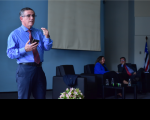 Homem em pé, vestido com camisa azul e gravata vermelha, usando óculos com lentes de grau. A mão direita ele está segurando o celular e a mão esquerda está fazendo gestos. Ao fundo, duas pessoas sentadas em poltronas de cor preta.
