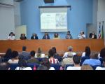 Foto de homens e mulheres sentadas em frente a uma grande de mesa, além de outras pessoas sentadas em cadeiras em um auditório. Na parede central do auditório, há um projetor. À direita, as bandeiras do Brasil e do Maranhão.