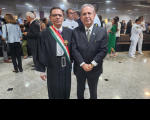 Dois homens posicionados em pé, posando para foto, ambos usando óculos de grau.  O homem da esquerda usa toga na cor preta e faixa transpassada listrada nas cores verde, branca e vermelha, com a comenda. Ao fundo, algumas pessoas em pé circulando pelo ambiente.