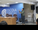 Foto de uma mulher em pé que está gesticulando com as mãos. Ela usa roupas nas cores verde claro e pretas e está com o cabelo preso num coque. A mulher está ao lado de uma mesa e cadeiras e próxima a uma parede azul onde está escrito Escola Judicial na cor branca.