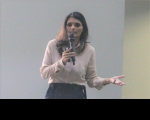 Foto de mulher clara, com cabelos escuros, usando blusa de mangas compridas em tom de rosa claro e saia preta. Ela fala ao microfone, que segura com a mão direita, e gesticula com a mão esquerda. Ao fundo, vê-se parede em tons claros.