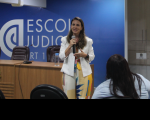 Foto de uma mulher que está em pé, falando ao microfone. Ela veste macacão colorido e um blazer branco. Na frente dela, há uma mulher sentada. Atrás dela, há uma mesa com cadeiras, e uma parede azul onde tem uma logomarca e está escrito Escola Judicial na cor branca.