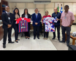 Foto de três mulheres e quatro homens em pé em uma sala. Duas mulheres seguram uma camisa com listras verticais e uma parte lisa, nas cores vermelha, azul, branca e preta; e um homem e outra mulher seguram uma camisa com fundo branco e figuras nas cores azul, vermelha e preta.