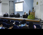 Quatro pessoas fazendo a abertura de um seminário dentro de um auditório e uma intérprete de libras. 