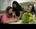 Foto de três mulheres preparando ikebanas com um arranjo de flores amarelas em primeiro plano.