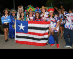 Grupo de oito mulheres posando para foto e segurando a bandeira do Estado do Maranhão (nove listras horizontais intercaladas em três cores: vermelho, branco e preto), e no lado superior esquerdo da bandeira, um quadrado azul com uma estrela na cor branca). 