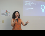 Foto de uma mulher branca, de cabelos encaracolados, usando uma blusa laranja, falando ao microfone, que segura com a mão direita, e gesticula com a mão esquerda. Ela está à frente de uma parede branca, na qual se vê um cartaz à sua direita e uma projeção de slides à sua esquerda.