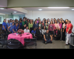 Foto de mulheres e homens em uma sala ampla. Parte das pessoas está em pé, outras estão agachadas e um homem está sentado. No ambiente há uma mesa coberta por uma toalha rosa. Ainda sobre a mesa um arranjo floral na cor rosa, além de celulares.