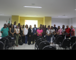 Foto de várias mulheres e vários homens que estão em pé num auditório. Estão em frente a uma parede amarela e branca.