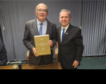 Dois homens dentro de uma sala com cortinas de persianas ao fundo, posando para foto. Ambos usando óculos de grau, vestidos com terno escuro e gravata na cor azul. O homem da esquerda segura um livro amarelo em suas mãos.