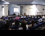 Foto de várias pessoas (homens e mulheres) sentadas num auditório participando de seminário; no destaque, palestrante. Na parede central do auditório, um projetor com as informações do evento. À direita, o brasão do Judiciário na parte superior da parede.