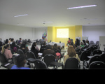 Foto de mulheres e homens num auditório. Todos estão sentados e, em frente a uma parede amarela, há uma mesa coberta por uma toalha branca, e duas mulheres à direita que estão debatendo, observadas por um homem, que está no lado esquerdo, e por pessoas que estão na plateia. No ambiente, há também bandeiras oficiais e banners.