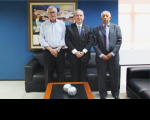 Três homens posando para foto, dentro de uma sala com parede de fundo na cor azul, mesa de centro em madeira com objetos decorativos em formato esférico..