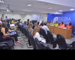 Foto de homens e mulheres sentados em um auditório, que estão assistindo a uma mulher que está em pé e falando ao microfone, em frente a uma parede azul, onde está escrito Escola Judicial. A mulher tem cabelos compridos e escuros e está vestindo um macacão verde. 