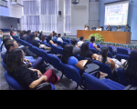Foto de várias pessoas (homens e mulheres) num auditório. Parte das pessoas está sentada em cadeiras azuis e a outra parte está sentada em cadeiras colocadas junto a uma ampla mesa, no palco do auditório. Atrás do palco, uma tela de projeção. As paredes do auditório são pintadas nas cores azul e cinza. Também tem no ambiente aparelhos de ar condicionado e cortinas de persianas.