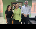 Foto de duas mulheres e um homem, todos de pele clara. A mulher ao centro usa blazer verde e saia preta. A da esquerda usa calca e blusa pretas. O homem, posicionado à direita, usa calça preta e camisa branca com grid preto. Todos sorriem em pose para a foto e estão de pé à frente de uma parede verde com um quadro artístico colorido centralizado.