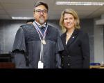 Duas pessoas posando para foto dentro de um auditório. Um homem à esquerda, vestido com toga, e uma mulher vestida com blaser preto.