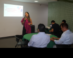 Foto de uma mulher clara, de cabelos longos e escuros, em pé, falando ao microfone, que segura com a mão direita. Ela usa blusa rosa e calça preta e está  à frente de uma parede branca, onde se projeta uma apresentação de slides. Na plateia, veem-se pessoas de costas, em mesas circulares, assistindo à palestra.