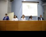 Mesa de honra composta por cinco pessoas, três mulheres ao centro e dois homens nas extremidades. A foto foi tirada dentro de um auditório com paredes azul e branca.