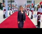 Foto de uma mulher branca, cabelos curtos e claros, usando óculos escuro, vestido e blaser preto, de pé sobre tapete vermelho na entrada do STF e ladeado por guardas usando o fardamento oficial dos Dragões da Independência.