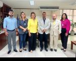 Seis pessoas posando para foto em pé, lado a lado, sendo três mulheres e três homens, dentro de uma sala com paredes brancas.