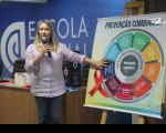 Foto de uma mulher em um auditório com parede de fundo azul, branca, cabelos loiros, vestida com camisa listrada colorida e uma calça jeans. Com a mão esquerda ela segura o microfone próximo a boca e com a direita, aponta para banner, com gráfico colorido, escrito PREVENÇÃO COMBINADA.