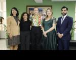 Cinco pessoas posando para foto, sendo quatro mulheres e um homem, dentro de uma sala. Ao fundo, parede verde com quadro colorido.