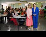 À frente, duas mulheres posando para foto, dentro de uma sala. Ao fundo, mulheres sentadas ao redor de mesas redondas e outras muheres em pé.