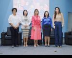 Foto de cinco mulheres de pé, lado a lado, posando e sorrindo para a câmera. Atrás delas, veem-se poltronas pretas e uma tela de projeção em uma parede azul, exibindo ilustração colorida.