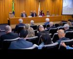 Foto de um auditório, com paredes ao fundo revestidas com madeira. Mesa solene composta por três pessoas, dois homens e uma mulher (ao centro). 