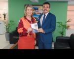 Duas pessoas posando para a foto, sendo uma mulher e um homem, dentro de uma sala, com parede de fundo esverdeada. Amulher usa saiade cor laranja e camisa social em tons terrosos. O homem usa terno e calça social em tom de azul. Eles seguram, ao centro, a Carta de Serviços da AGU-MA.