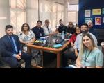 Várias pessoas, homes e mulheres, sentados em torno de uma mesa, posando para a foto. Eles estão dentro de uma sala.