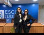 Duas pessoas posando para foto, dentro de uma sala. Ao fundo, parede azul escrita com letras brancas ESCOLA JUDICIAL.