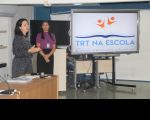 Foto de duas mulheres de pé em auditório, fazendo apresentação. Elas estão posicionadas à esquerda e, enquanto a de vestido preto fala, a de blusa roxa a observa. À direita, vê-se tela de projeção com a logomarca do Programa TRT na Escola.