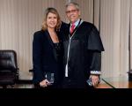 Duas pessoas posando para foto dentro de uma sala. Uma mulher, com vestido e blazer preto, segurando na mão uma bolsa preta, e um homem vestido com toga preta.