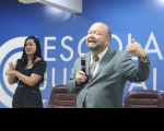 Foto de um homem branco e calvo, sorrindo e falando ao microfone, que segura com a mão direita. Ele usa terno cinza e gesticula com a mão esquerda. Em segundo plano, vê-se uma mulher branca de cabelos longos e pretos, usando roupa preta, gesticulando ao interpretar em LIBRAS a fala do homem. Ao fundo, vê-se parede azul com o texto ESCOLA JUDICIAL escrito em letras brancas, e os espaldares de duas cadeiras marrons.