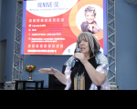 Foto de mulher branca, falando ao microfone, que segura com a mão esquerda e gesticula com a direita. Ao fundo, vê-se tela de projeção com arte da palestra.