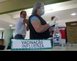 uma mulher, usando luvas com uma agulha na mão. À frente, placa escrita VACINAÇÃO INFLUENZA.