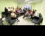 Grupo de pessoas posando para foto dentro de uma sala. O ambiente possui paredes de cor verde, cadeiras pretas e mesa de madeira.